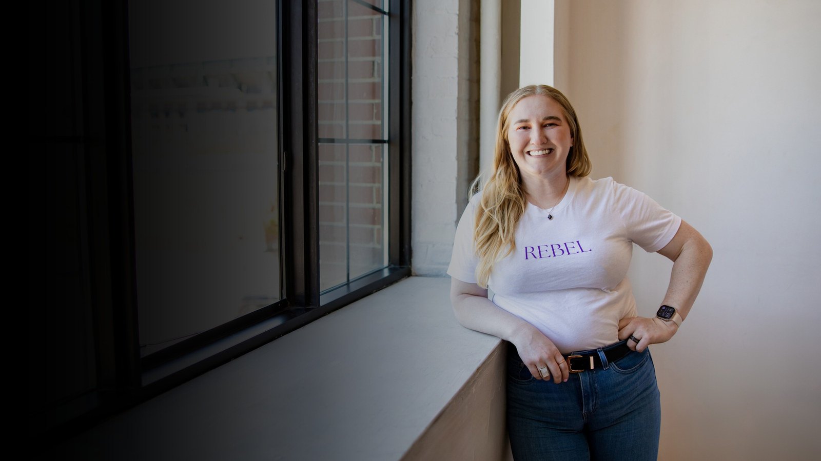 Jenica Norris smiling by a window, wearing a REBEL t-shirt